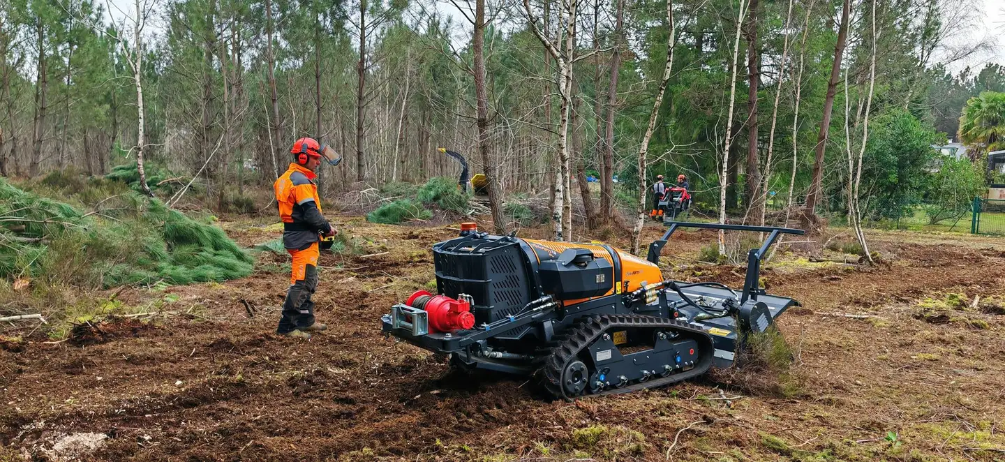 Terrain débroussaillé jusqu'à la lisière de la pinède, opérateur à la télécommande du broyeur, habitations en arrière-plan
